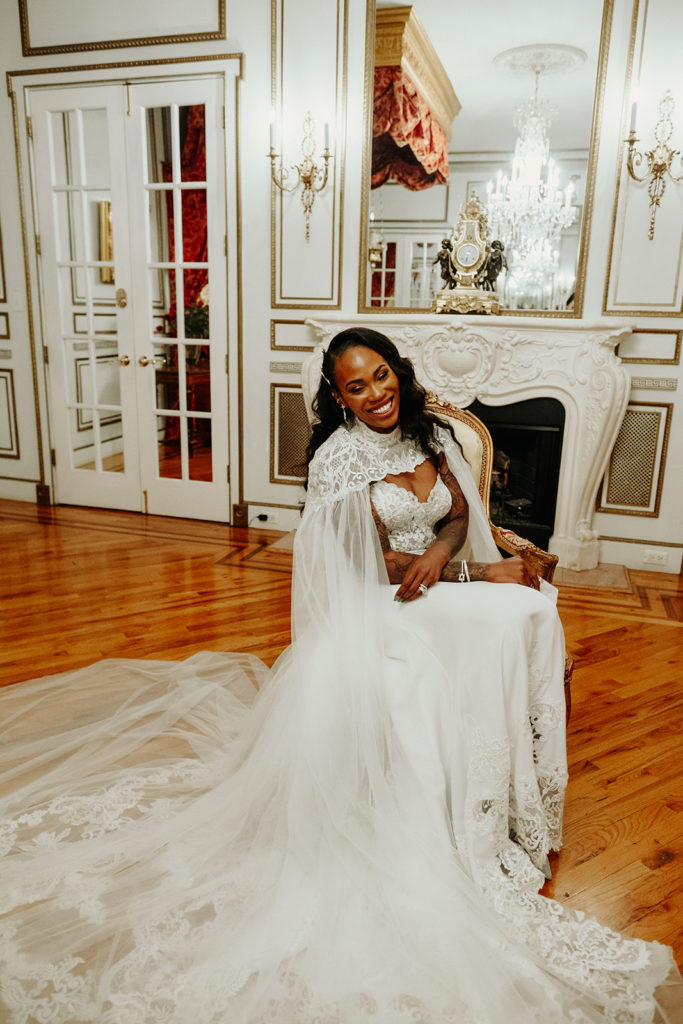 Bride wearing Calla Blanche Wedding Gown and Cape sits in Chair at James Ward Mansion Wedding
