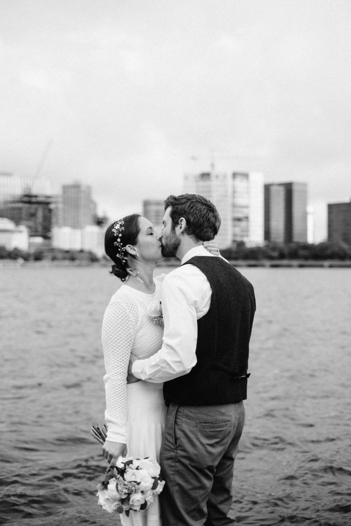 Bride and groom kiss along the charles river with cambridge skyline behind them