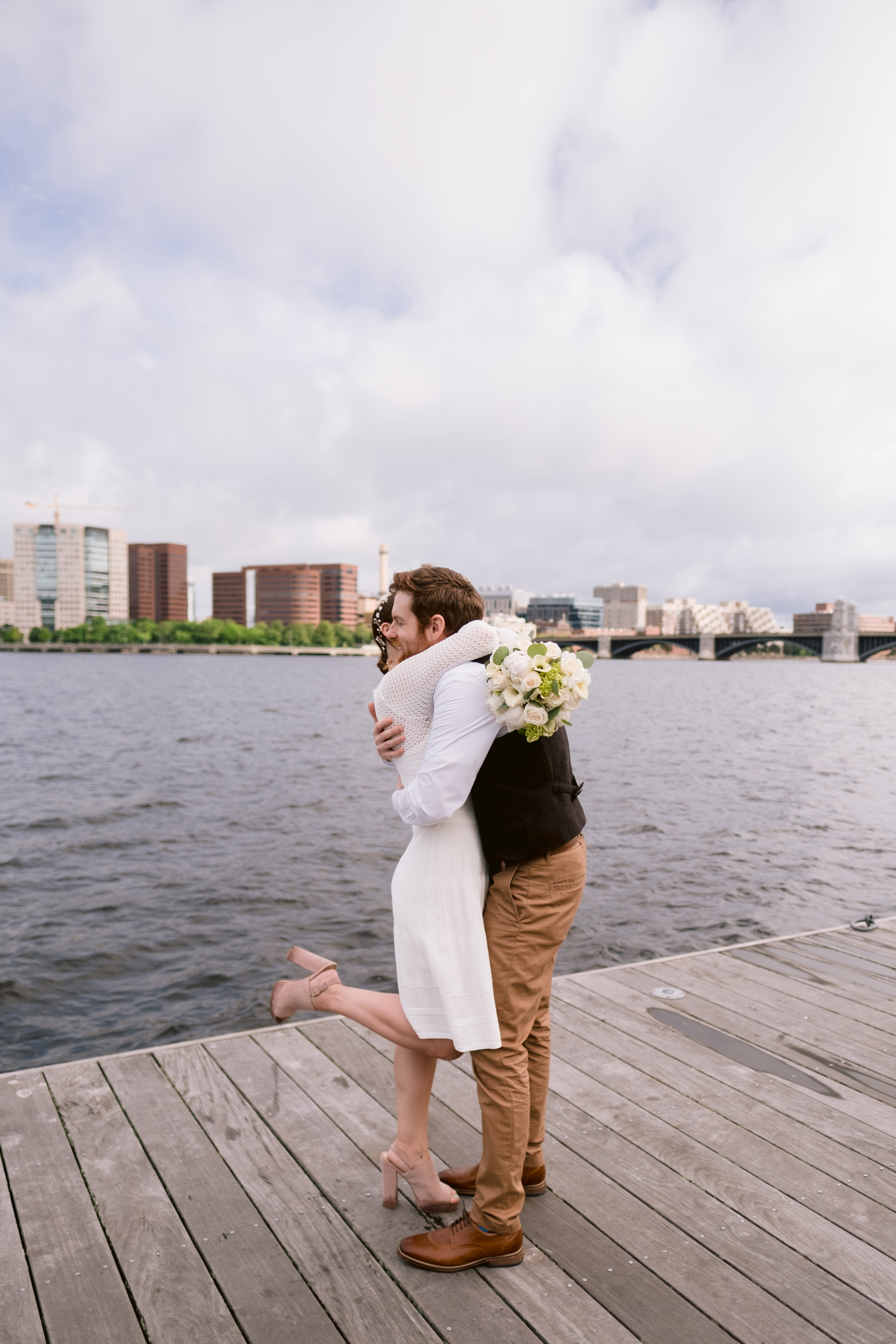 Bride and groom embrace along the charles river with cambridge skyline behind them during Boston Elopement