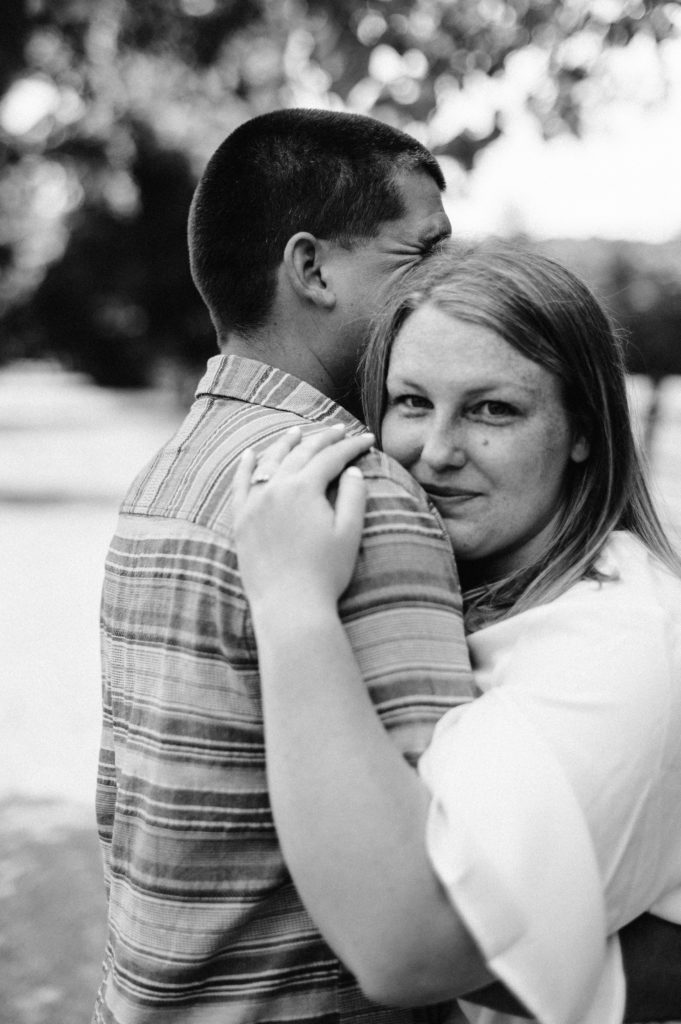 Black and white portrait of woman peering over man's shoulder with engagement ring on her hand