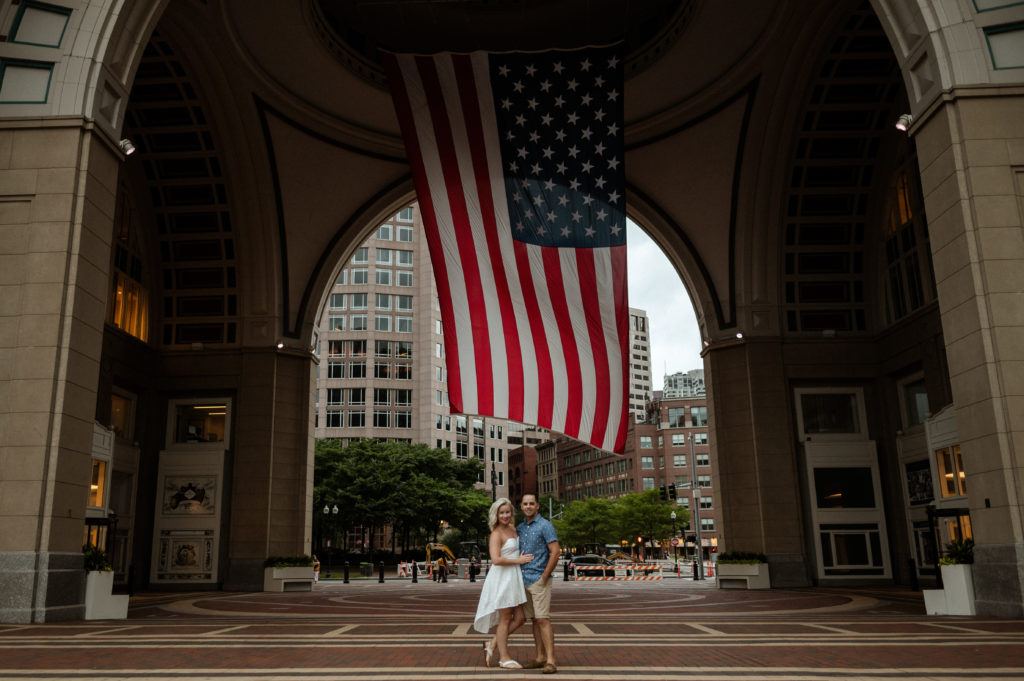 Couple stands in front of large american flag during Boston Seaport Engagement Session