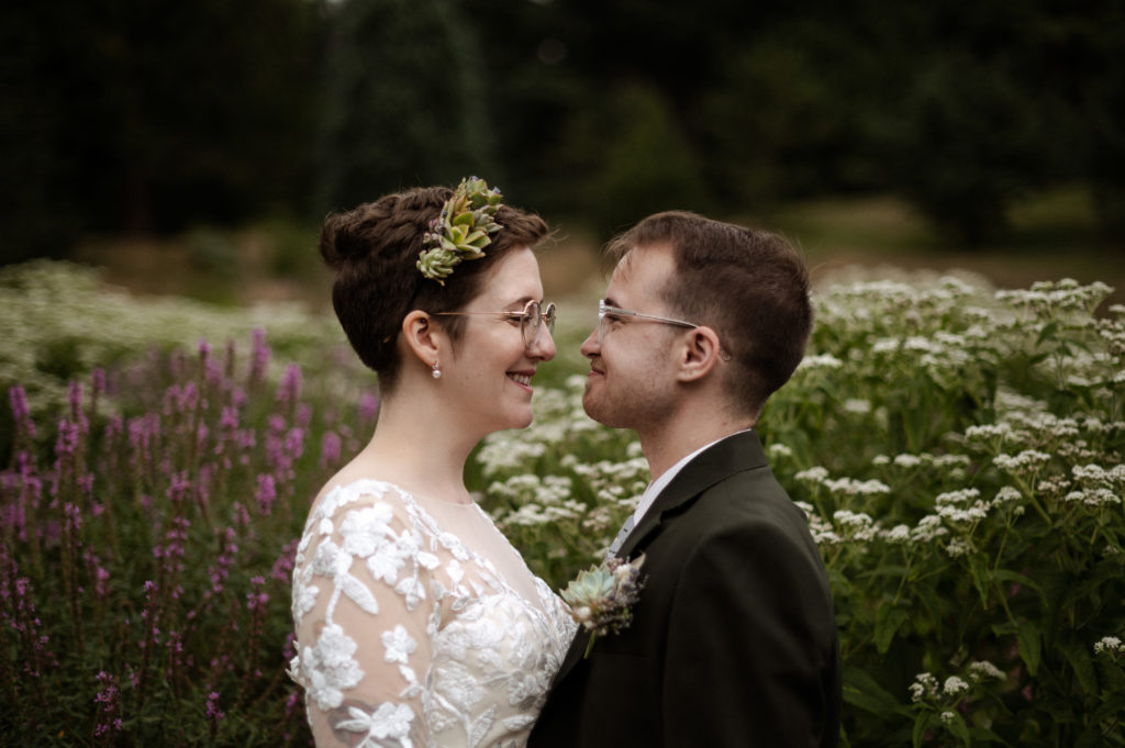 Bride and groom show of succulent hairpiece and boutonniere sit in wildflowers in Arnold arboretum during COVID 19 Elopement in Boston