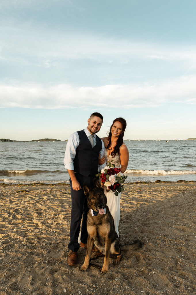 Bride and groom pose with their German Shepherd wearing a bowtie during their Boston Minimony on Wollaston Beach Quincy