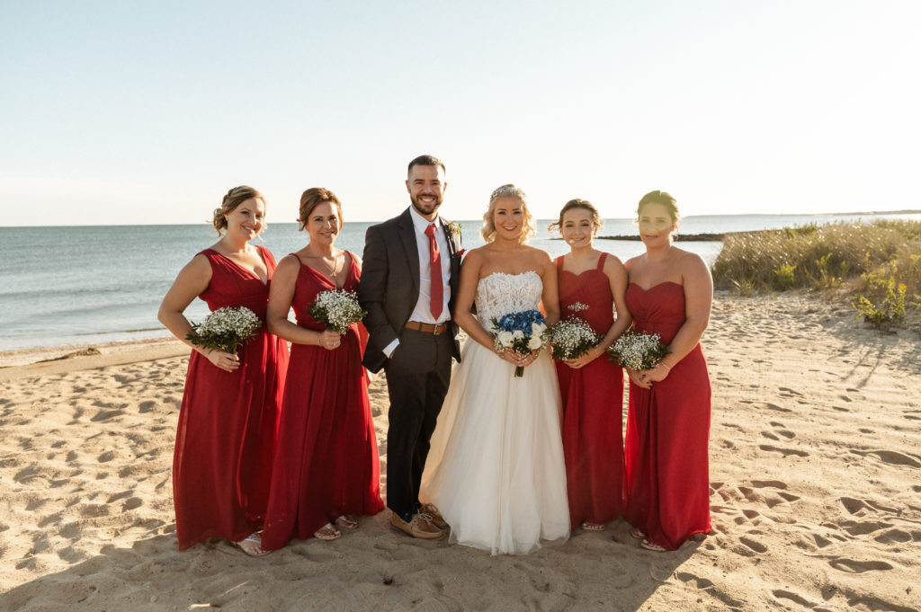 Bride with mixed gender bridal party and bridesmaids in red dresses on beach at The Lighthouse Inn at Cape Cod Wedding
