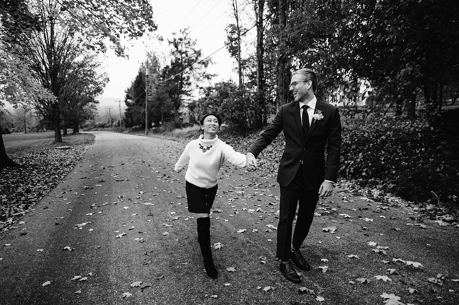 B&W Groom holding Bride's hand and leading down fallen leaf road