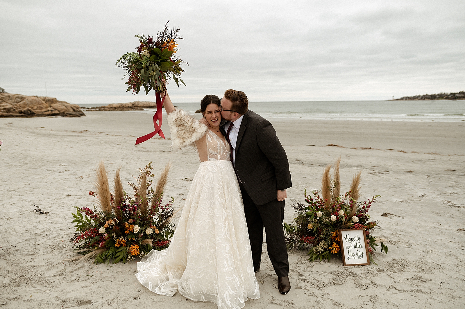 Bride lifting up bouquet with Groom kissing Bride on the cheek on the beach