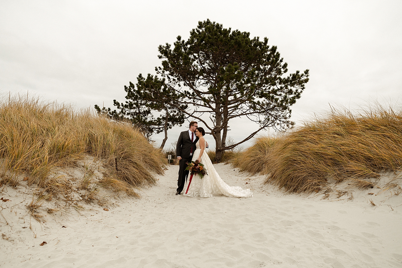 Wide shot of Groom holding Bride standing on sand with tree behind them