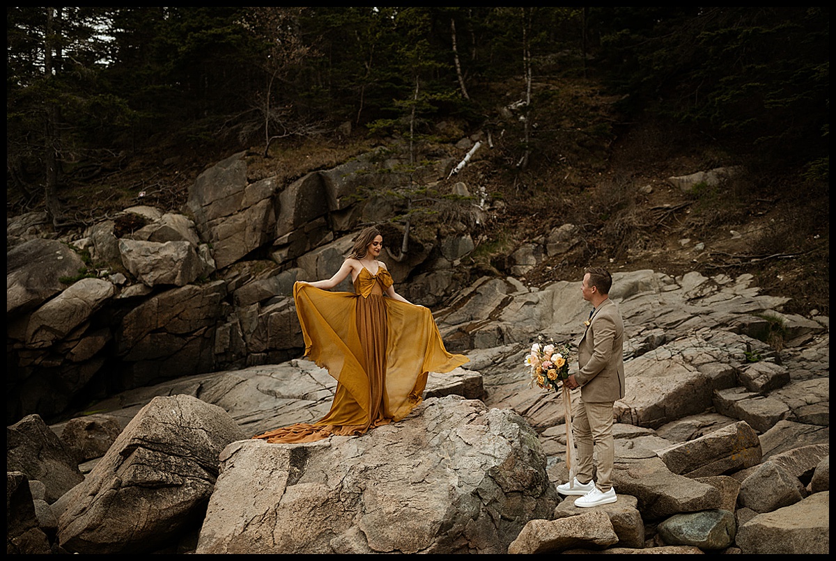 Bride in Golden Leanne Marshall Gown stands on rocks as groom looks at her with bouquet at Sand Beach Maine